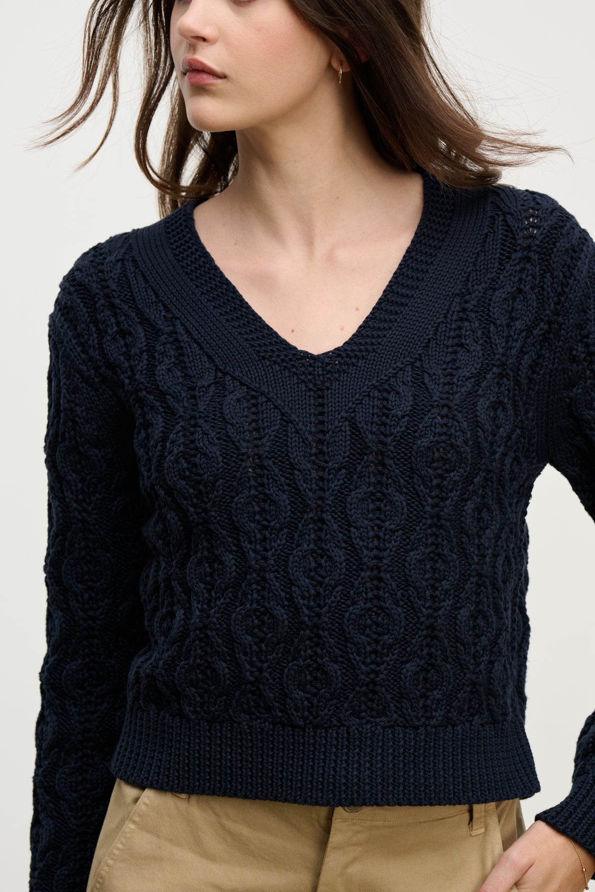   A woman wears the JOYCE SWEATER by Velvet by Graham & Spencer, a navy blue chunky Pima cotton cable knit with a boxy fit, paired with tan pants and shown from mid-thigh up against a light background. 