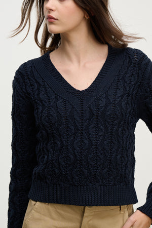 A woman wears the JOYCE SWEATER by Velvet by Graham & Spencer, a navy blue chunky Pima cotton cable knit with a boxy fit, paired with tan pants and shown from mid-thigh up against a light background.
