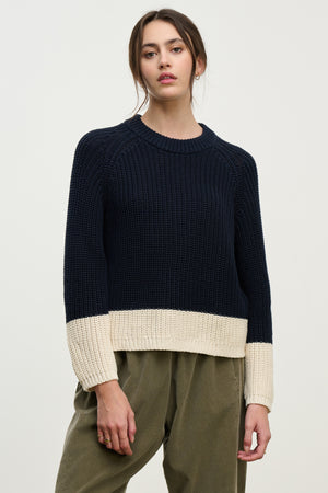 A woman stands against a plain background wearing the SHAY SWEATER by Velvet by Graham & Spencer in navy Pima cotton with cream cuffs and hem, paired with olive green pants.