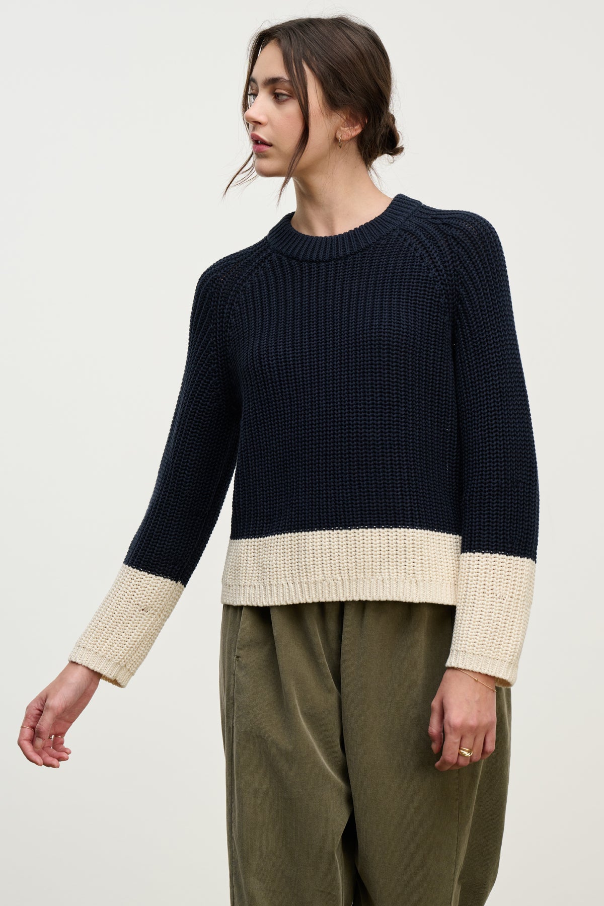 A person wears the SHAY SWEATER by Velvet by Graham & Spencer—an oversized color block crewneck in navy blue and cream—with olive green pants, standing against a plain light background.-39800388452545