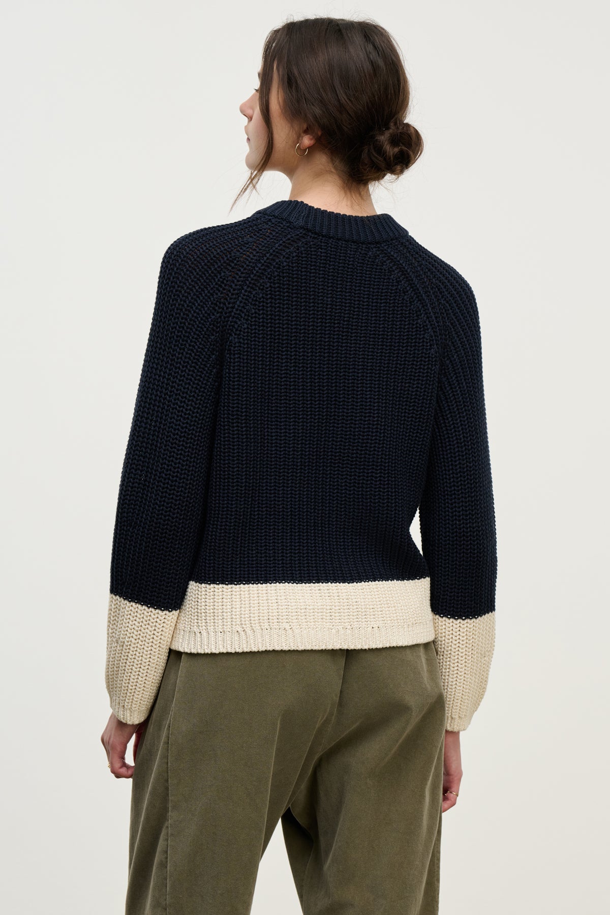 A person with brown hair in a low bun is shown from the back wearing the SHAY SWEATER by Velvet by Graham & Spencer, a navy blue color block style with beige cuffs and hem, paired with olive green pants.-39800388387009