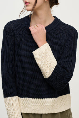 A woman models the SHAY SWEATER by Velvet by Graham & Spencer, featuring a navy blue color block design with cream cuffs and hem, holding her hand near her neck.