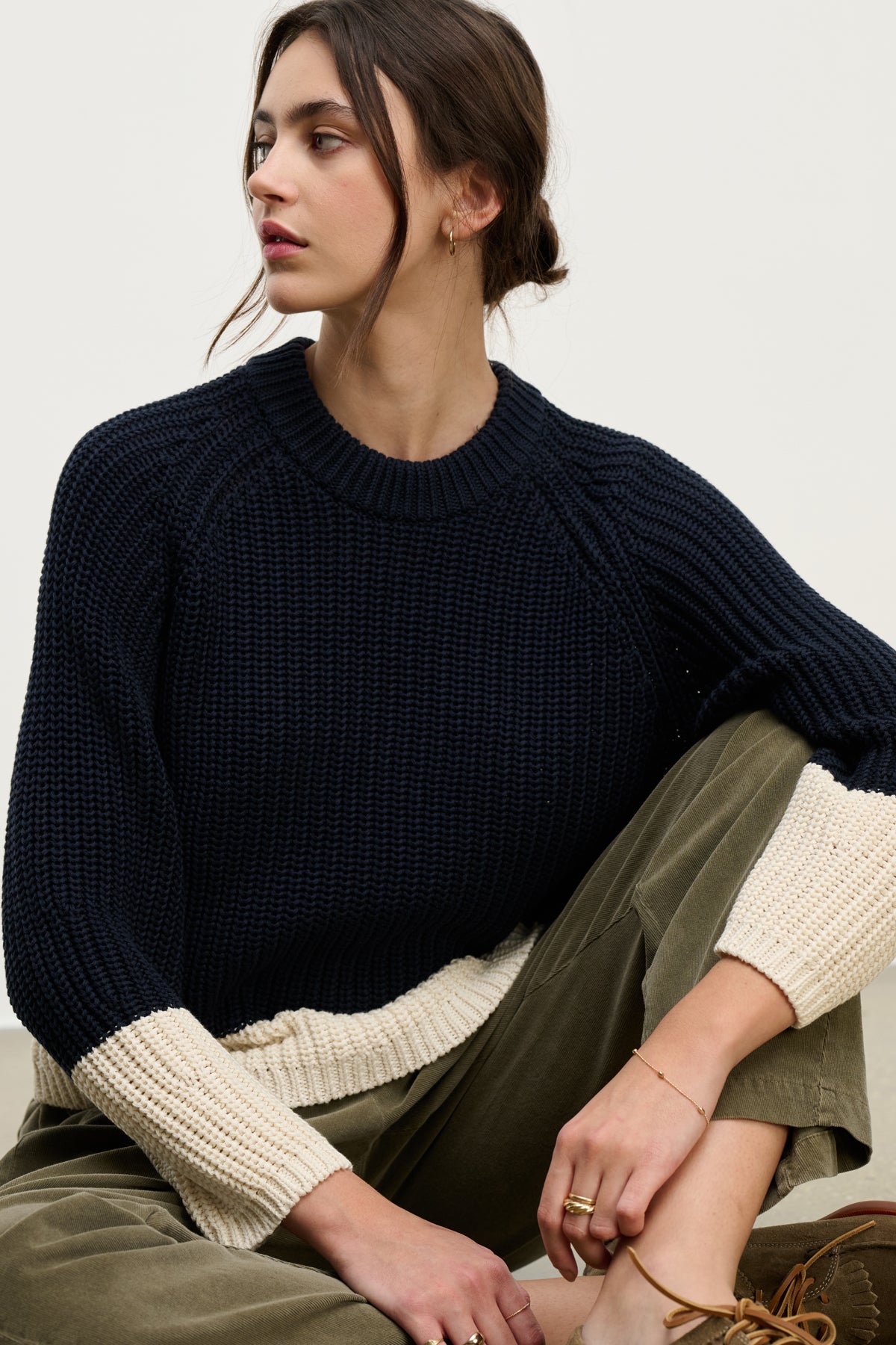   A woman with dark hair in a low bun wears the Velvet by Graham & Spencer SHAY SWEATER, a navy blue color block style with cream cuffs, paired with olive green pants. She is seated and looking to the side. 