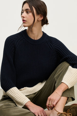 A woman with dark hair in a low bun wears the Velvet by Graham & Spencer SHAY SWEATER, a navy blue color block style with cream cuffs, paired with olive green pants. She is seated and looking to the side.