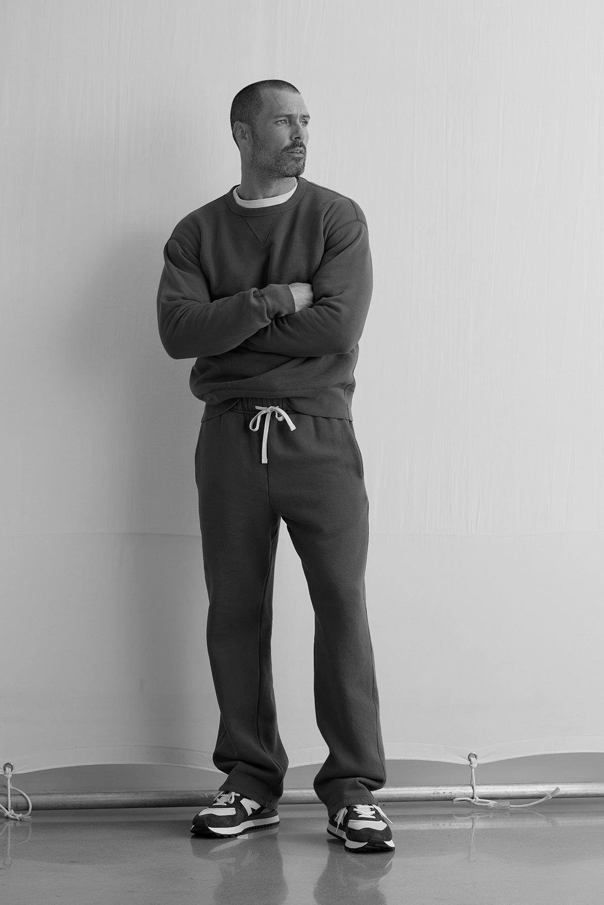 A man stands against a plain wall with arms crossed, wearing the Velvet by Graham & Spencer CILLIAN SWEATSHIRT, paired with drawstring sweatpants and sneakers.-40277059436737