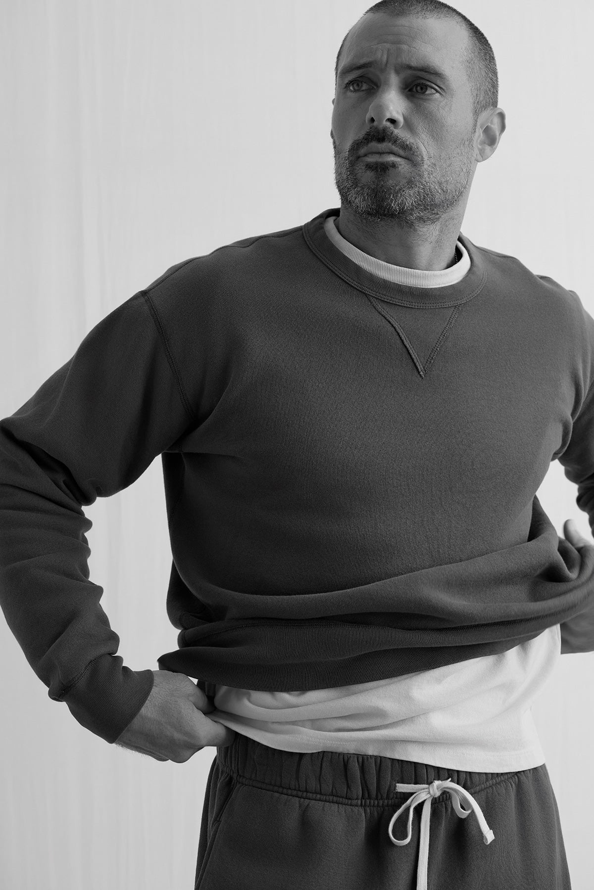 A man stands with one hand on his hip against a plain background, wearing the Velvet by Graham & Spencer CILLIAN SWEATSHIRT over a t-shirt and drawstring pants.
