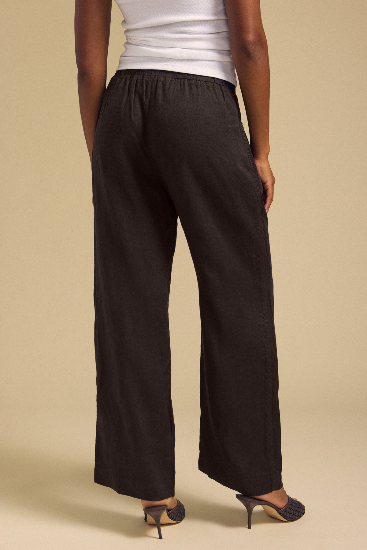 A person stands facing away, wearing a white sleeveless top paired with the CLAIRO LINEN PANT by Velvet by Graham & Spencer—black, wide-leg linen pants with a pull-on elastic waist—and black heeled shoes, set against a beige background.