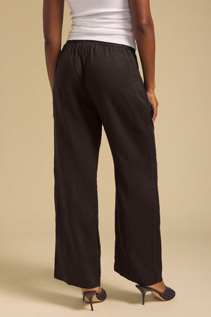 A person stands facing away, wearing a white sleeveless top paired with the CLAIRO LINEN PANT by Velvet by Graham & Spencer—black, wide-leg linen pants with a pull-on elastic waist—and black heeled shoes, set against a beige background.