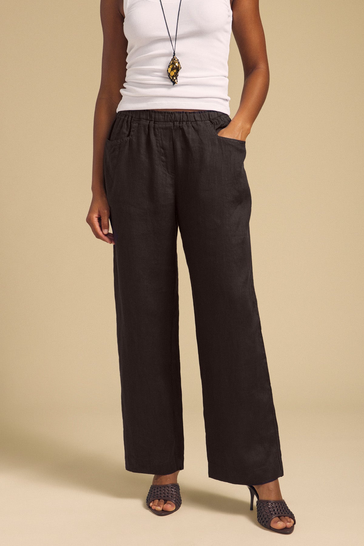 A person wearing a white sleeveless top, the CLAIRO LINEN PANT by Velvet by Graham & Spencer in black with an elastic waist and pockets, black heeled sandals, and a pendant necklace, standing against a beige background.