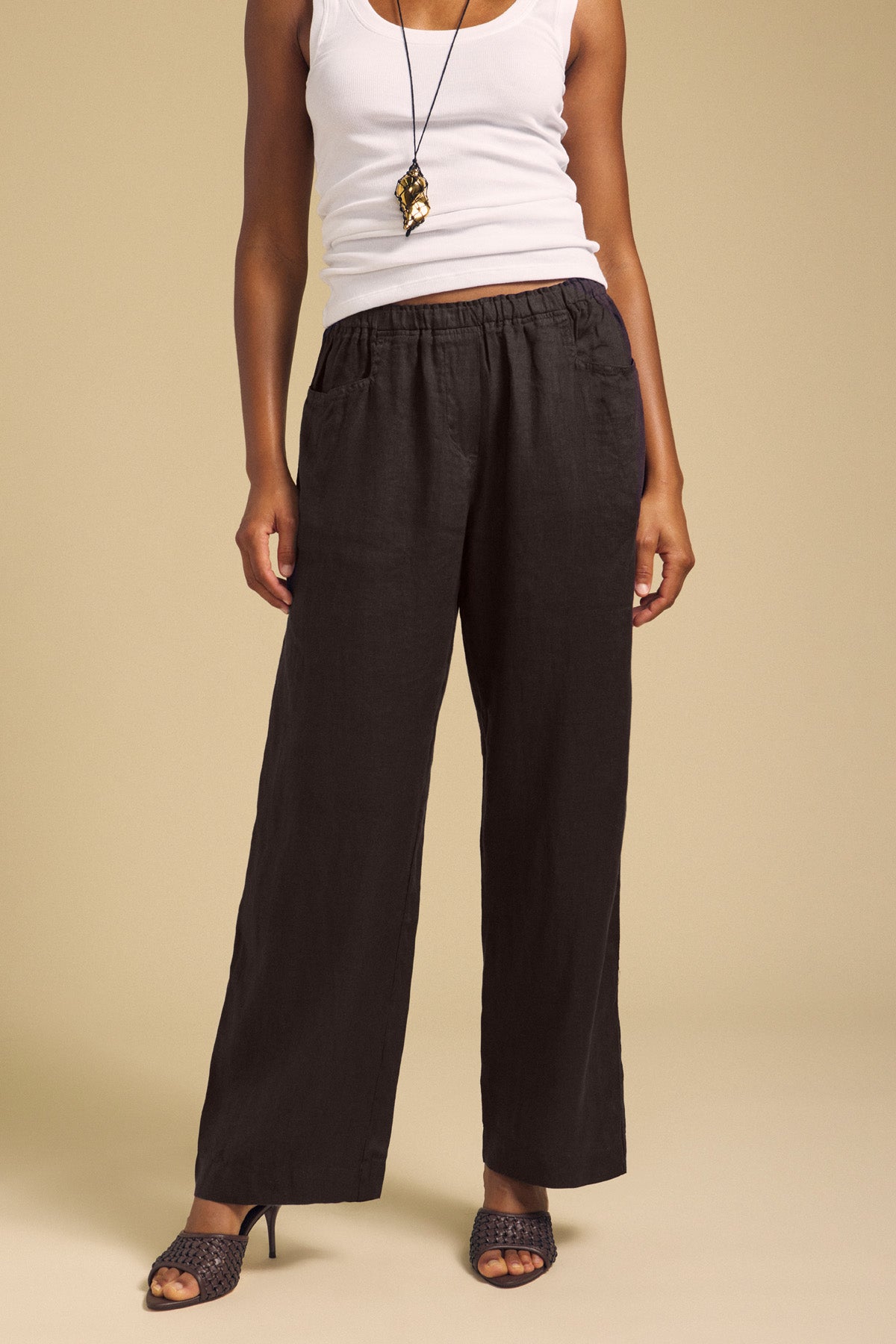 A person wearing a white tank top, the Velvet by Graham & Spencer CLAIRO LINEN PANT in black with an elastic waist, black heeled sandals, and a pendant necklace stands against a beige background.-41997210321089