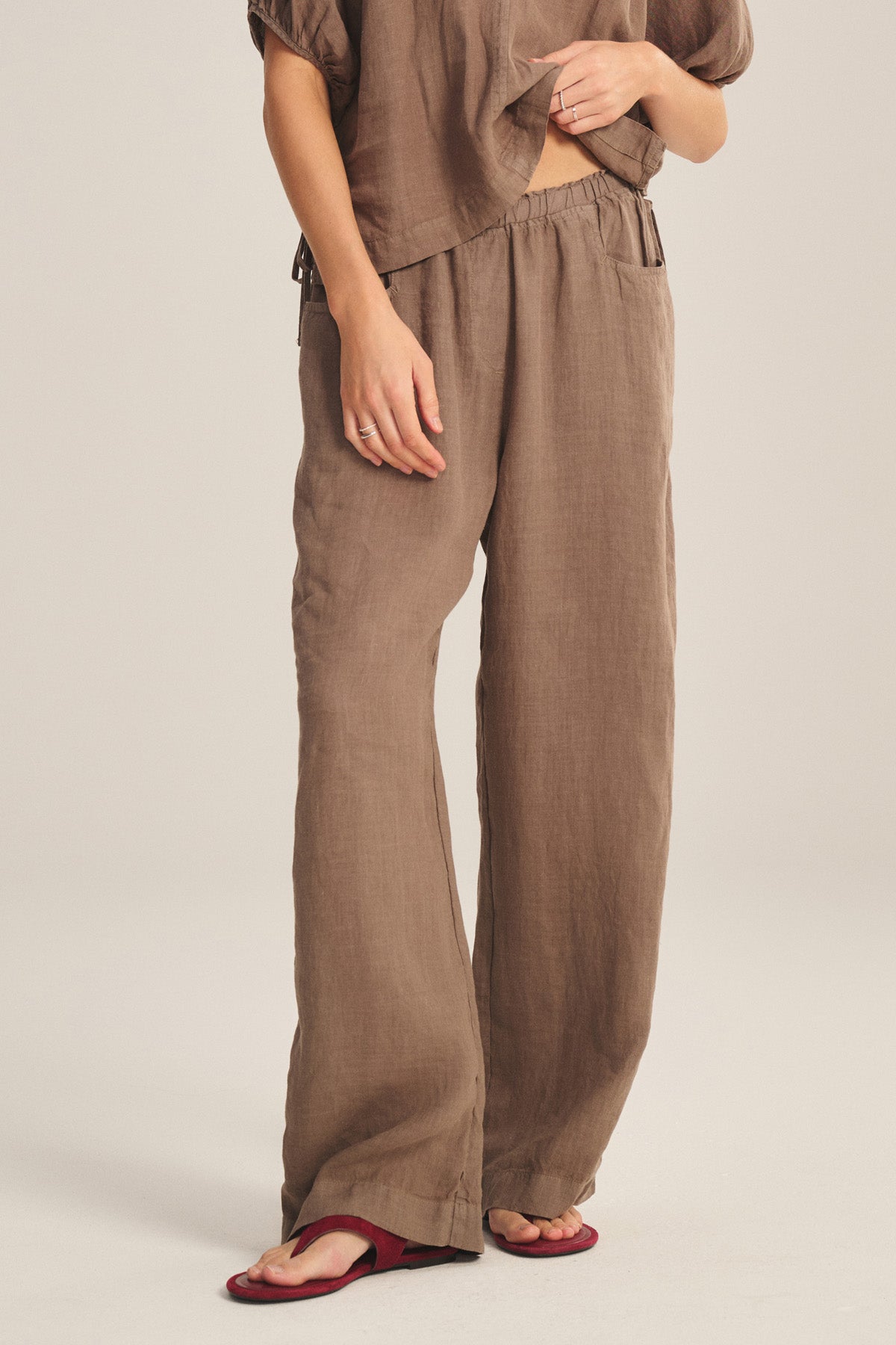 A person models the Velvet by Graham & Spencer CLAIRO LINEN PANT in brown—featuring a loose, wide-leg silhouette and elastic waist—with a matching top and red flat sandals against a plain light background; face not visible.