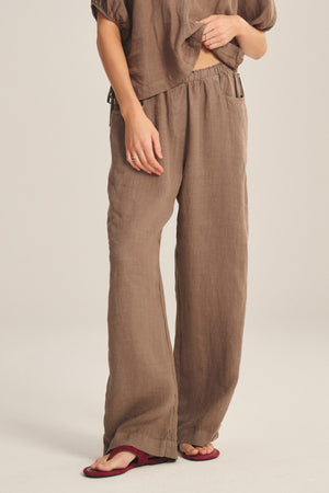 A person models the Velvet by Graham & Spencer CLAIRO LINEN PANT in brown—featuring a loose, wide-leg silhouette and elastic waist—with a matching top and red flat sandals against a plain light background; face not visible.