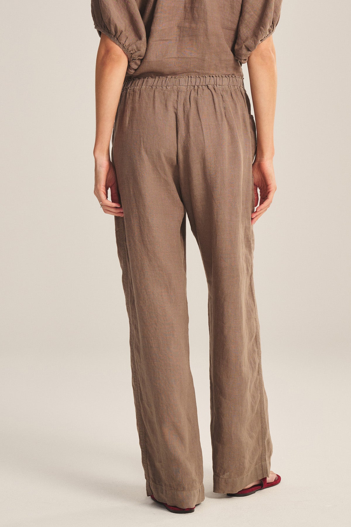 A person faces away, wearing the CLAIRO LINEN PANT by Velvet by Graham & Spencer—loose-fitting wide-leg brown linen pants with an elastic waist and a matching short-sleeve top, set against a plain background.
