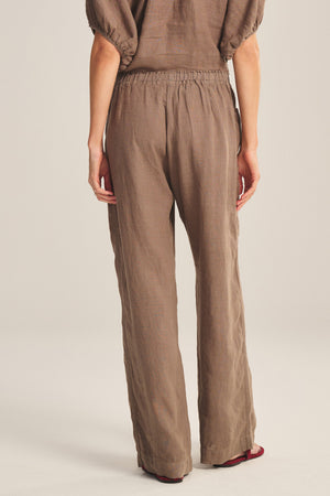 A person faces away, wearing the CLAIRO LINEN PANT by Velvet by Graham & Spencer—loose-fitting wide-leg brown linen pants with an elastic waist and a matching short-sleeve top, set against a plain background.