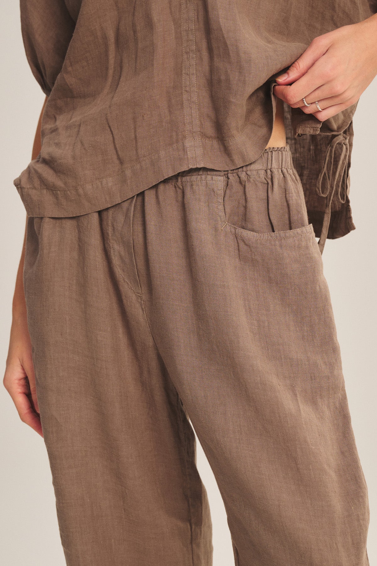 Someone wearing the CLAIRO LINEN PANT by Velvet by Graham & Spencer—a lightweight, loose-fitting linen pant with an elastic waist and side pocket—stands with one hand near their waist.