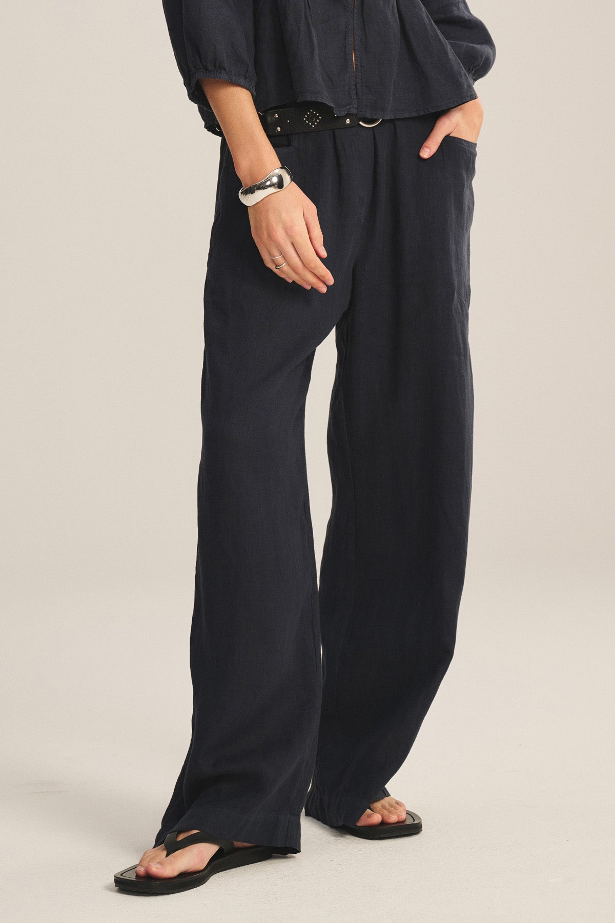 Wearing Velvet by Graham & Spencer's CLAIRO LINEN PANT, this person sports relaxed wide-leg linen pants with an elastic waist, paired with a black belt, black sandals, and a rolled-sleeve black long-sleeve top, hand in pocket.