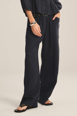 Wearing Velvet by Graham & Spencer's CLAIRO LINEN PANT, this person sports relaxed wide-leg linen pants with an elastic waist, paired with a black belt, black sandals, and a rolled-sleeve black long-sleeve top, hand in pocket.