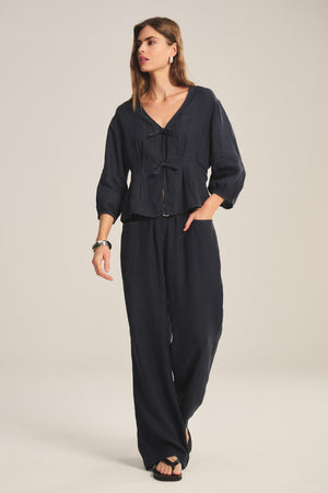 A woman stands against a plain light background wearing Velvet by Graham & Spencer’s CLAIRO LINEN PANT, a wide-leg, lightweight linen pant with an elastic waist, paired with a loose-fitting dark navy tie-front blouse.