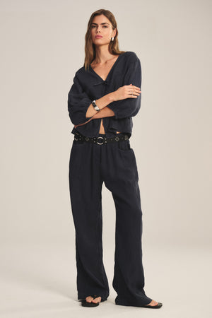 A woman stands with arms crossed, wearing Velvet by Graham & Spencer’s CLAIRO LINEN PANT—wide-leg, lightweight linen pants with an elastic waist—paired with a loose dark navy blouse and accessorized with a black belt and bracelet.