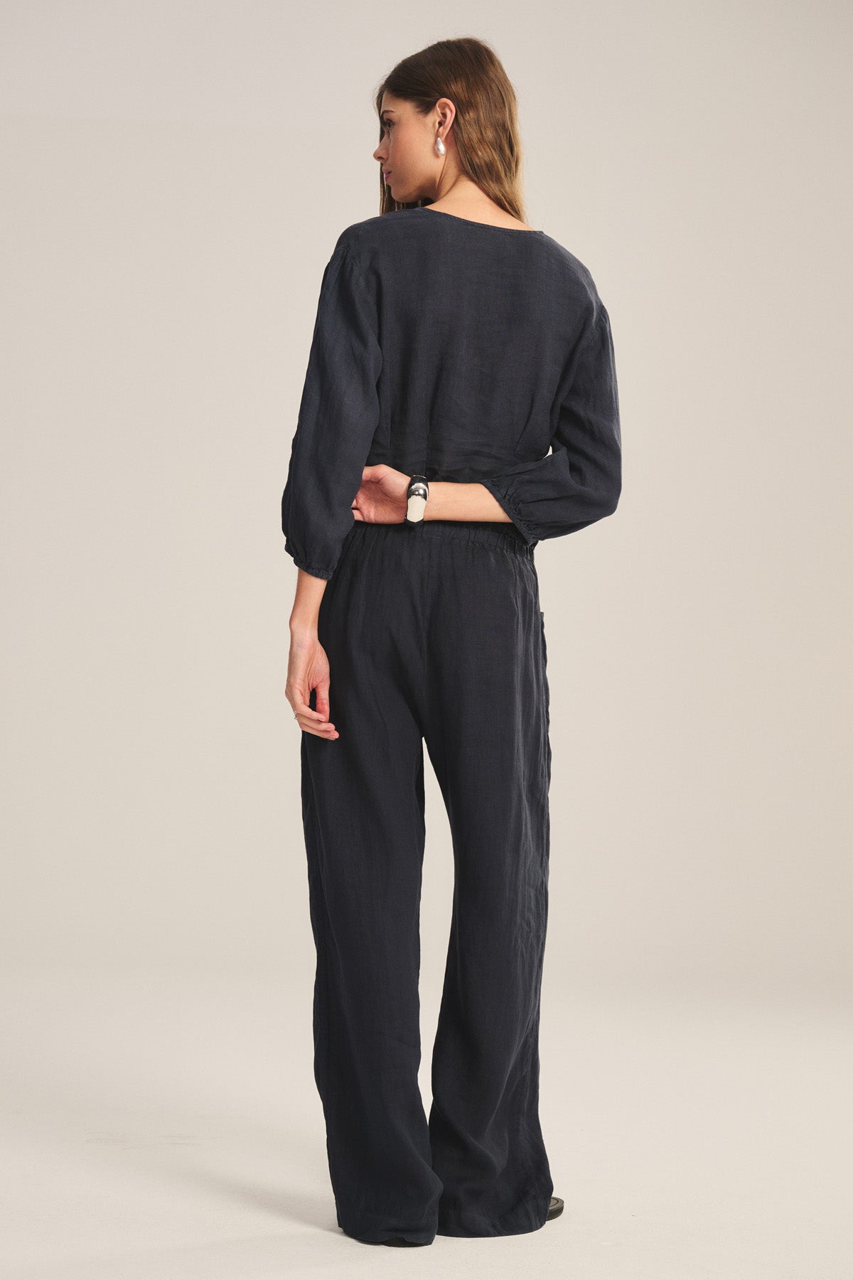 A woman stands facing away, wearing Velvet by Graham & Spencer's loose-fitting CLAIRO LINEN PANT in black, paired with a wristwatch, her left hand resting on her lower back.-41081234718913
