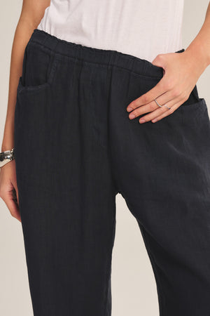 A person wears the CLAIRO LINEN PANT by Velvet by Graham & Spencer—wide-leg, lightweight linen pants with an elastic waist—paired with a white shirt, hand in pocket, showcasing a silver ring and bracelet.