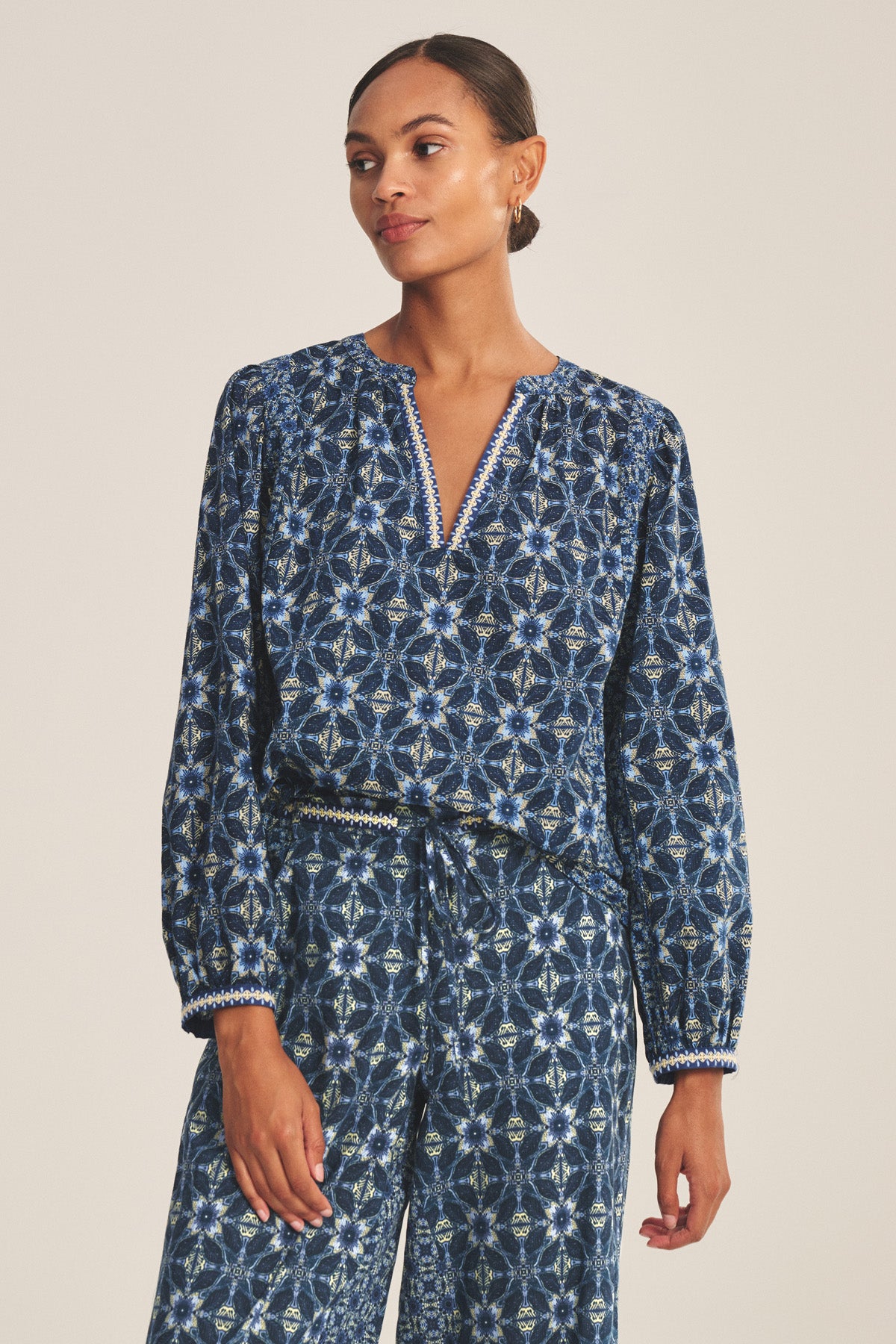 A person wearing the CLAUDIE TOP by Velvet by Graham & Spencer—a long-sleeved, printed cotton cambric top with a V-neck—paired with matching blue patterned trousers, stands against a plain background.-41008182591681