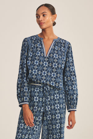 A person wearing the CLAUDIE TOP by Velvet by Graham & Spencer—a long-sleeved, printed cotton cambric top with a V-neck—paired with matching blue patterned trousers, stands against a plain background.