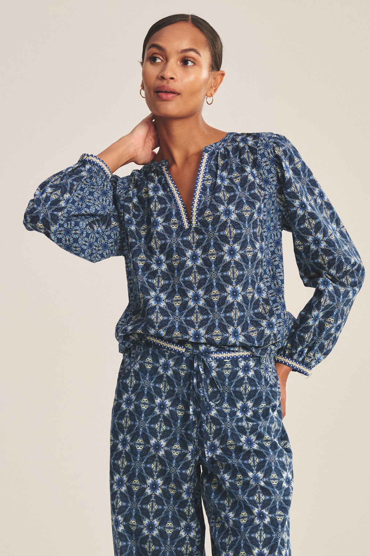 A woman wears the CLAUDIE TOP by Velvet by Graham & Spencer, a matching printed cotton cambric top and pants with a blue geometric pattern, standing with one hand on her neck while looking to the side.-41008182558913