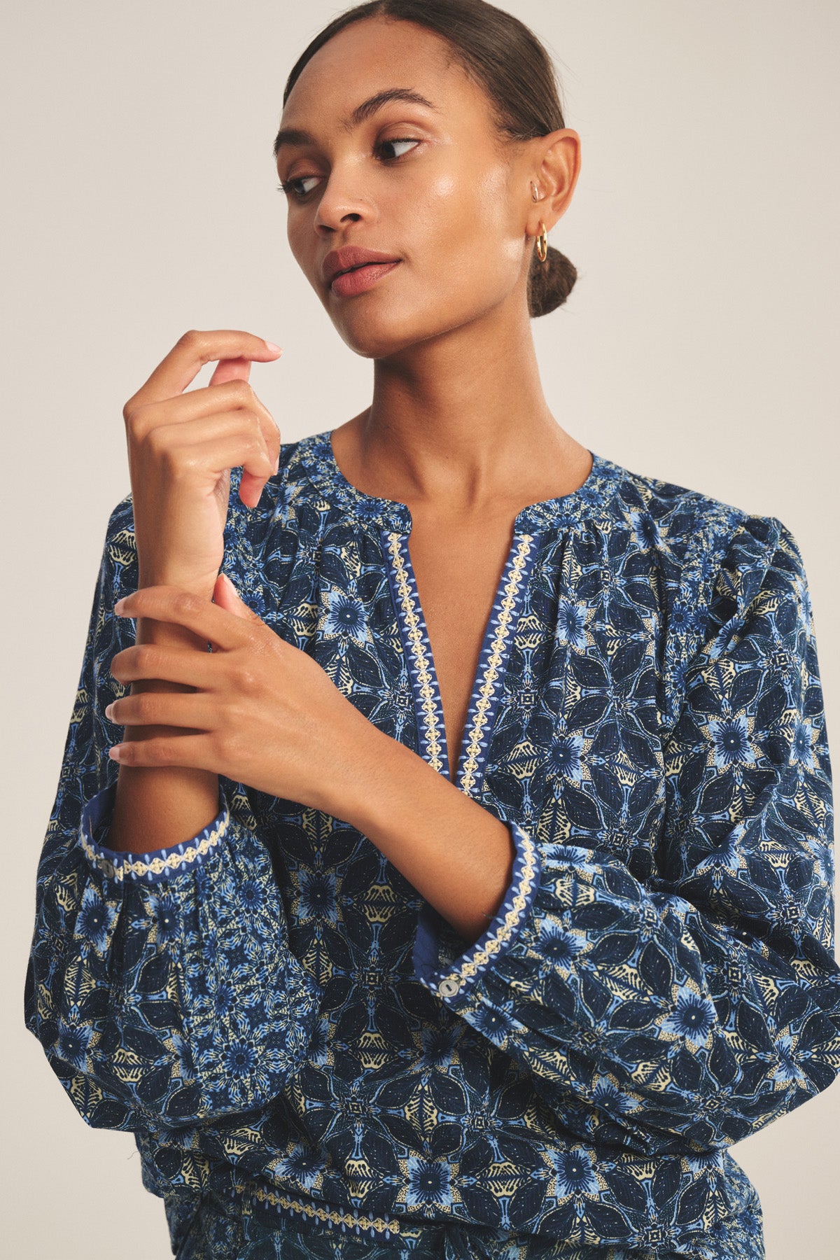 A woman wears the CLAUDIE TOP by Velvet by Graham & Spencer, a blue printed cotton cambric style with a notched neckline, as she looks to the side and touches her hand.