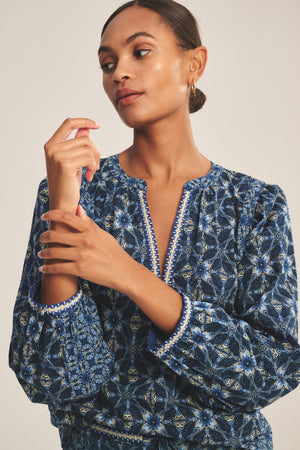 A woman wears the CLAUDIE TOP by Velvet by Graham & Spencer, a blue printed cotton cambric style with a notched neckline, as she looks to the side and touches her hand.