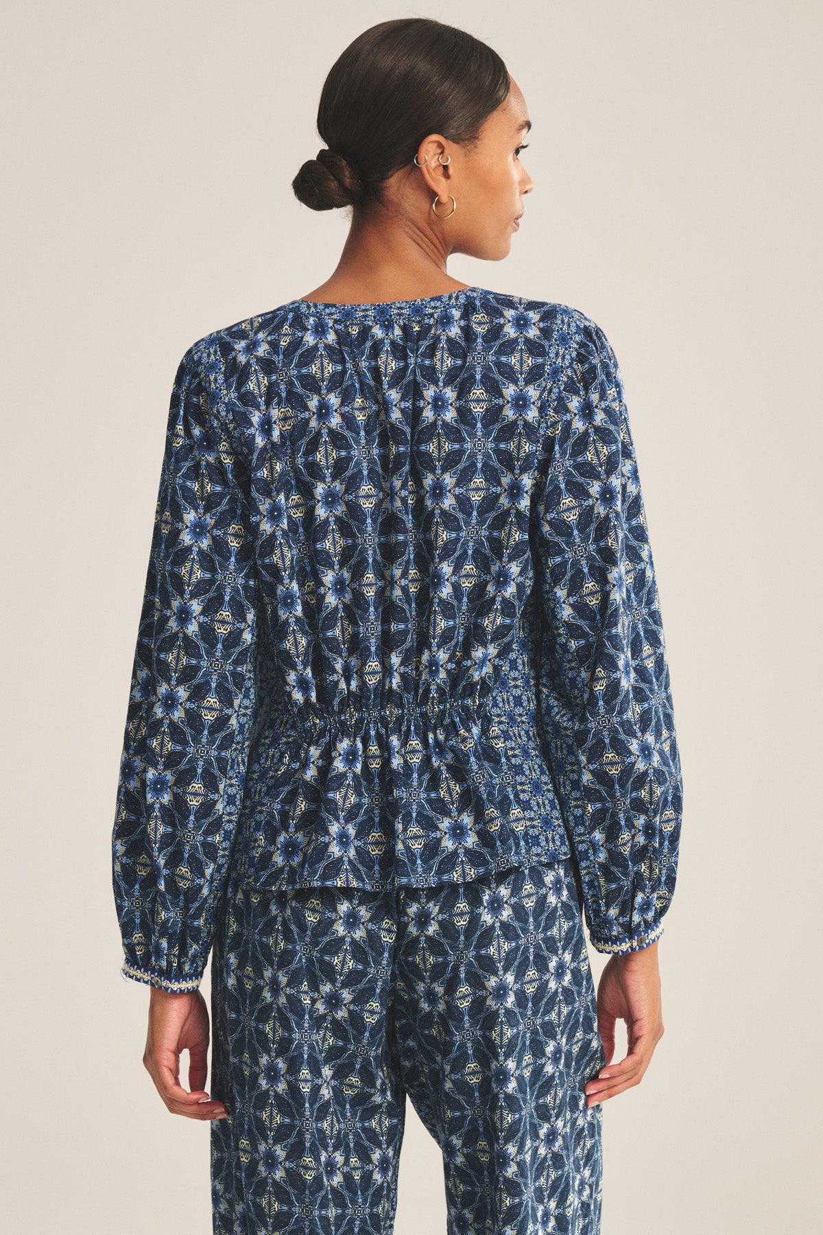 A woman stands facing away, wearing the CLAUDIE TOP by Velvet by Graham & Spencer—a printed cotton cambric top paired with matching blue patterned elastic-waist pants and a straight hem.-41008182395073