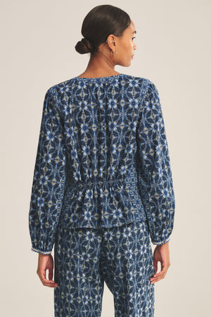 A woman stands facing away, wearing the CLAUDIE TOP by Velvet by Graham & Spencer—a printed cotton cambric top paired with matching blue patterned elastic-waist pants and a straight hem.