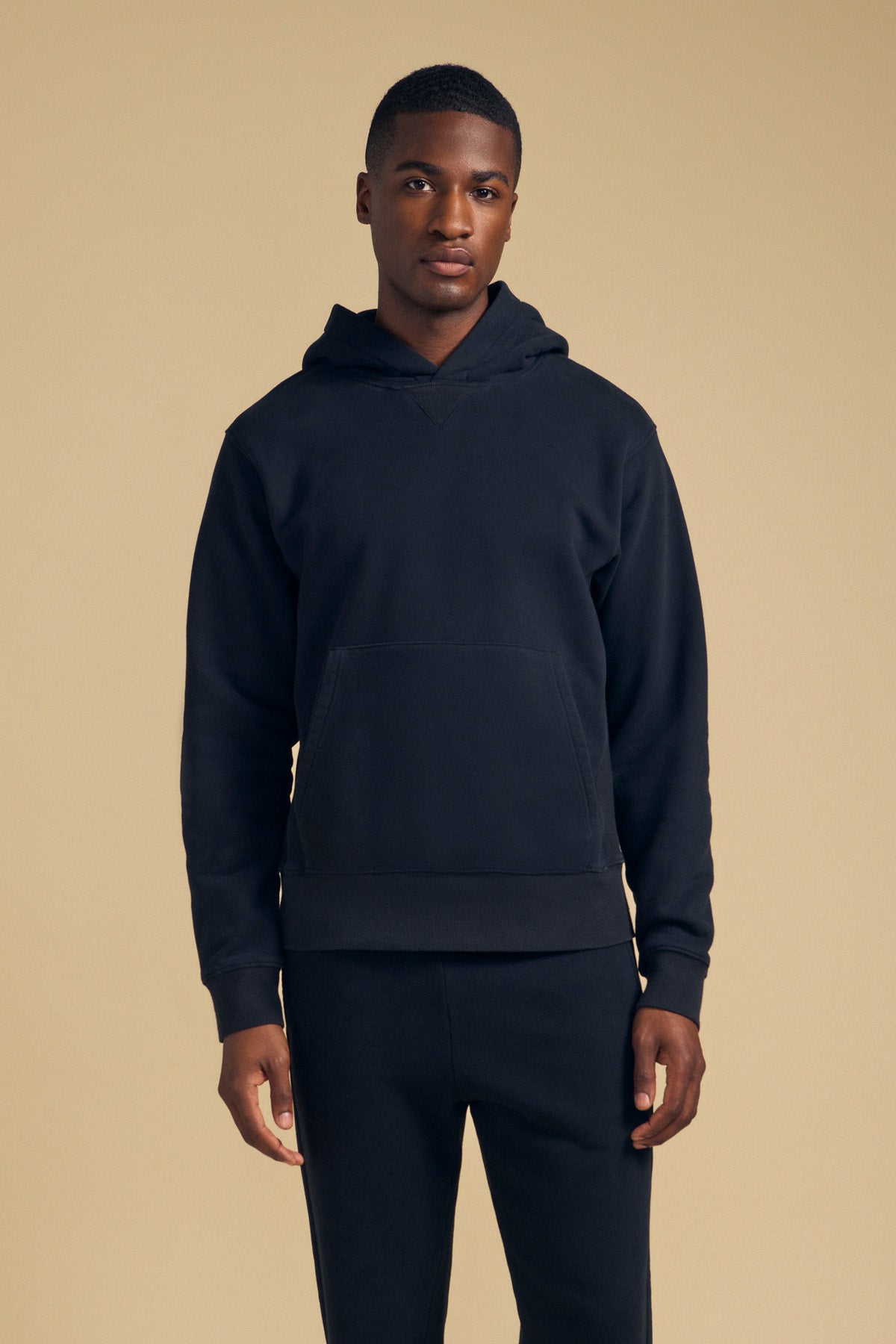 A man faces forward wearing the WES HOODIE and matching black pants from Velvet by Graham & Spencer against a plain beige background.-41702172098753