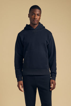 A man faces forward wearing the WES HOODIE and matching black pants from Velvet by Graham & Spencer against a plain beige background.