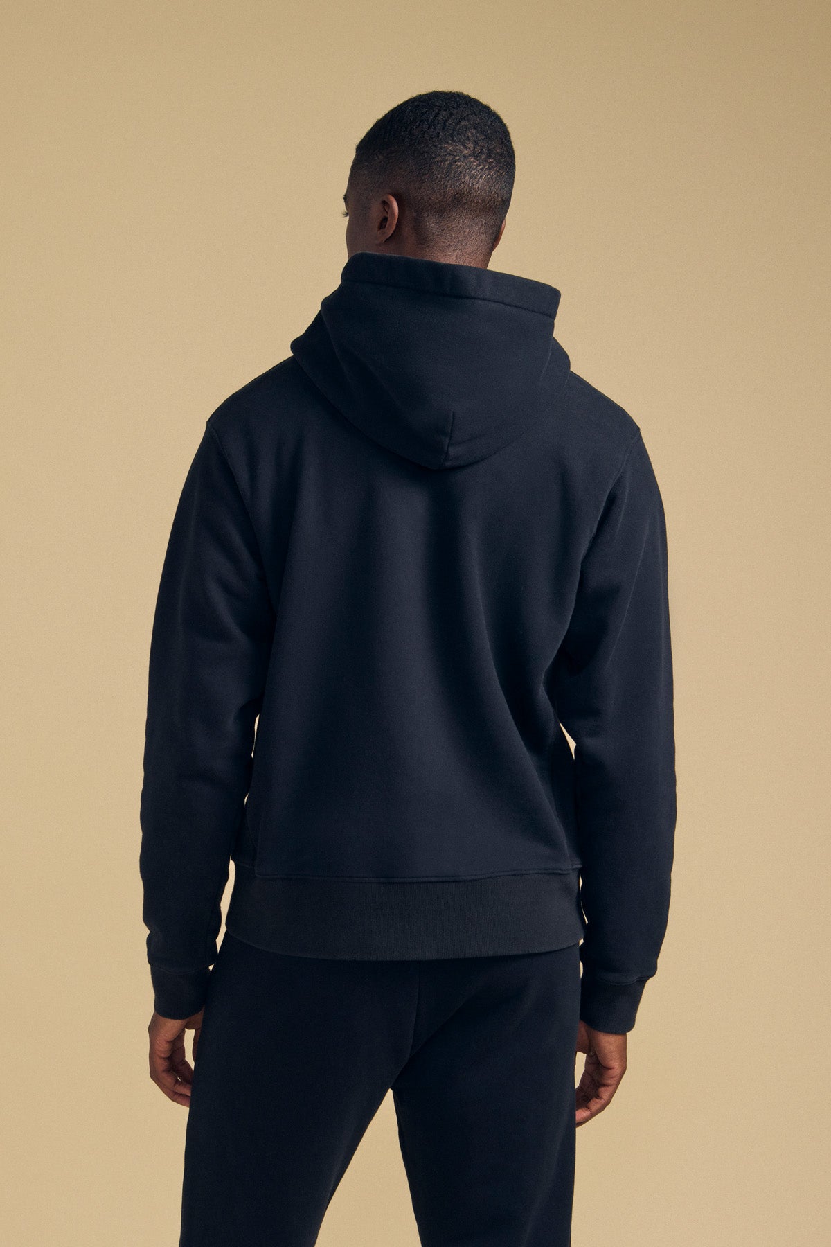 A person stands with their back to the camera, wearing the WES HOODIE in black by Velvet by Graham & Spencer and matching black pants, set against a beige background.-41702172033217