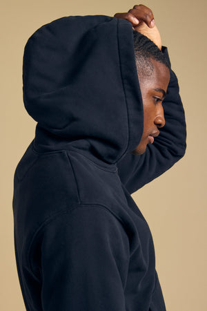 A person models the WES HOODIE by Velvet by Graham & Spencer in black, standing in profile against a beige background with one hand raised to the back of their head.