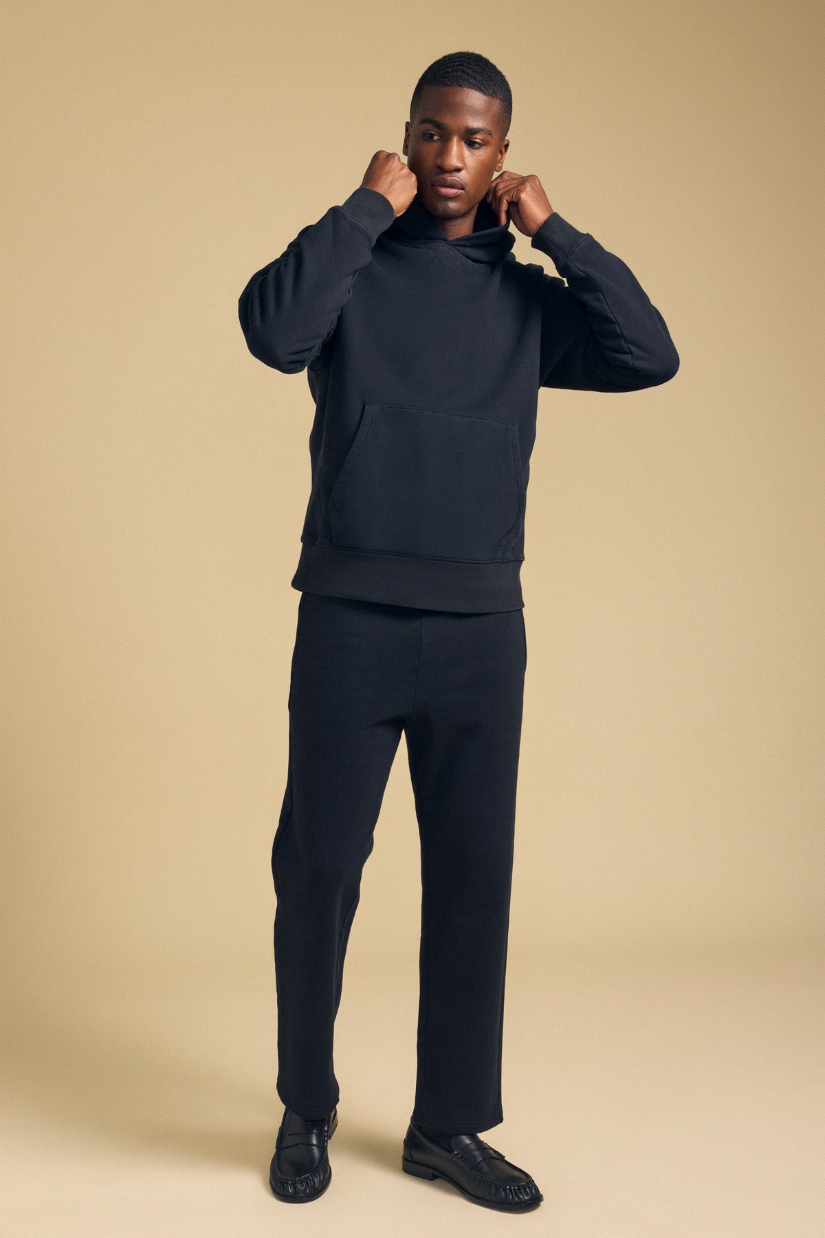 A person stands against a plain beige background wearing the WES HOODIE by Velvet by Graham & Spencer, paired with black pants and shoes, adjusting the hood with both hands.-41702171902145