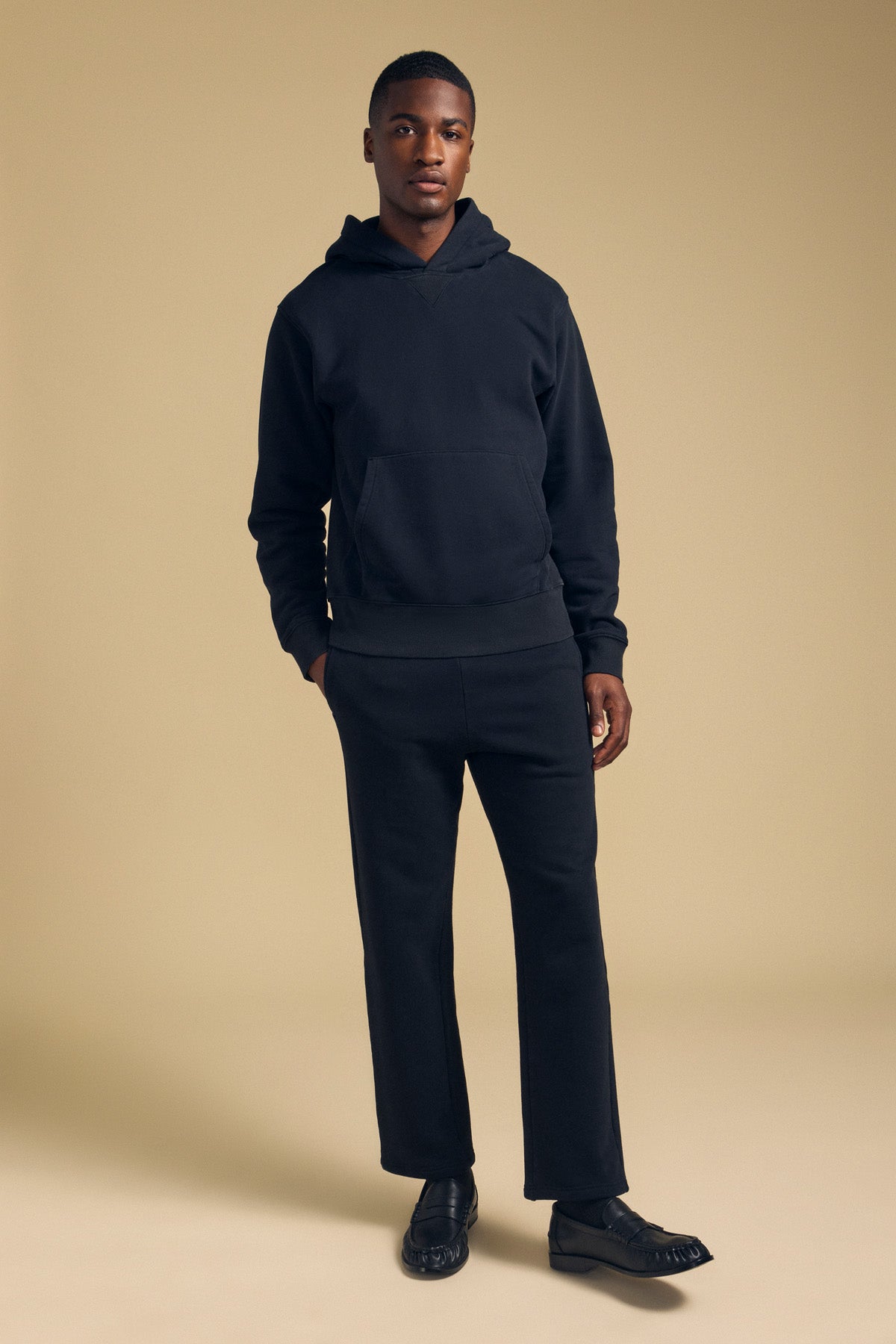 A man wearing a black hooded sweatshirt, black Velvet by Graham & Spencer HUDSON SWEATPANT relaxed fit sweatpants, and black loafers.
