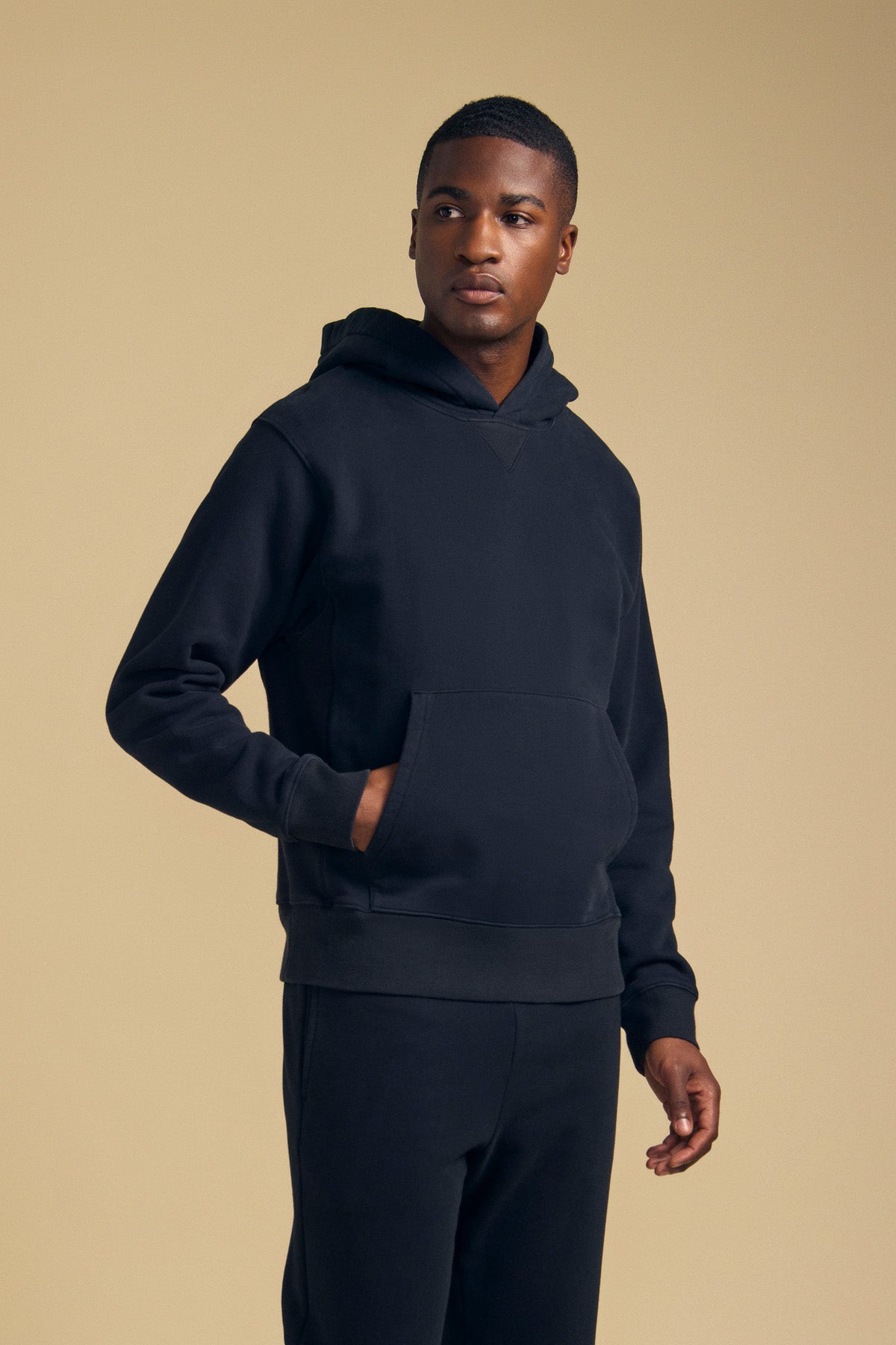 A man wears the WES HOODIE by Velvet by Graham & Spencer with matching pants, standing against a plain beige background and looking slightly to the side.-41702171803841