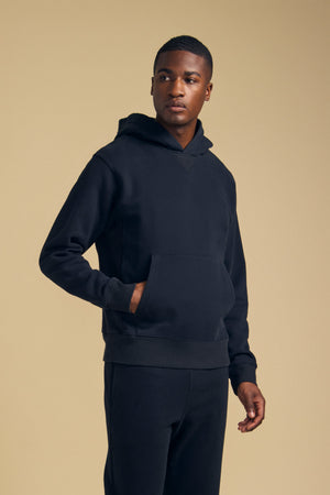 A man wears the WES HOODIE by Velvet by Graham & Spencer with matching pants, standing against a plain beige background and looking slightly to the side.