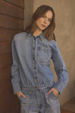 A woman with long brown hair stands indoors facing the camera, hands in her pockets, wearing the Velvet by Graham & Spencer CLEMENTINE DENIM SHIRT.