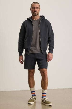 A man stands indoors against a plain background wearing the BRADFORD HOODIE by Velvet by Graham & Spencer, paired with a gray T-shirt, matching shorts, striped socks, and olive green sneakers.