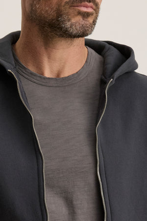 A close-up shows a man in a grey T-shirt and the unzipped BRADFORD HOODIE by Velvet by Graham & Spencer, revealing just the lower half of his face and his upper torso.