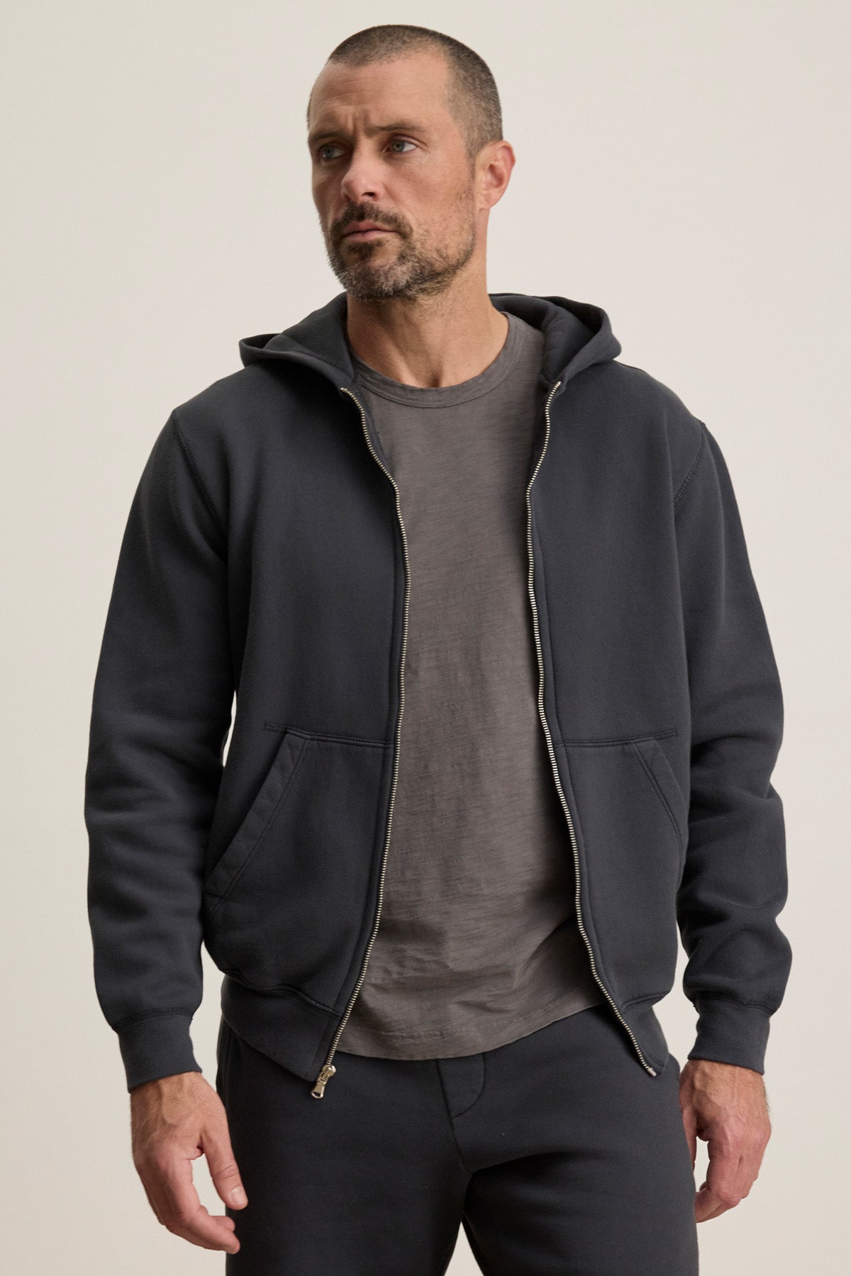 A man with short hair and a beard wears the Velvet by Graham & Spencer BRADFORD HOODIE, a durable full zip hoodie made from heavyweight fleece, over a gray t-shirt and matching pants, standing against a plain background.