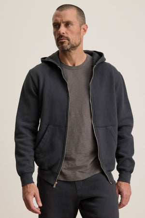 A man with short hair and a beard wears the Velvet by Graham & Spencer BRADFORD HOODIE, a durable full zip hoodie made from heavyweight fleece, over a gray t-shirt and matching pants, standing against a plain background.