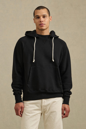 A person faces forward against a neutral background, wearing the BRODIE HOODIE by Velvet by Graham & Spencer—featuring a relaxed fit, drawstring hood, and white accents—paired with off-white pants.