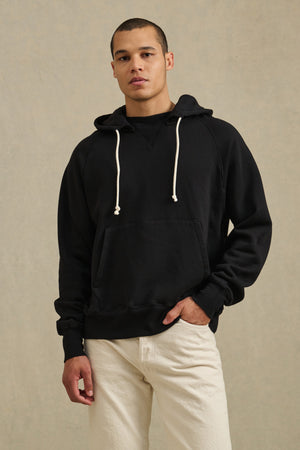 A man wearing the BRODIE HOODIE by Velvet by Graham & Spencer—a black relaxed fit hoodie with white drawstrings—and beige pants stands in front of a plain, light-colored background.