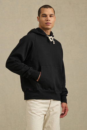 A person stands against a plain backdrop, wearing the BRODIE HOODIE by Velvet by Graham & Spencer—a black relaxed-fit hoodie with white drawstrings—and off-white pants, one hand in the hoodie pocket.
