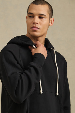 A man wearing the BRODIE HOODIE by Velvet by Graham & Spencer, a black relaxed fit style with white drawstrings, looks to the side against a neutral background.