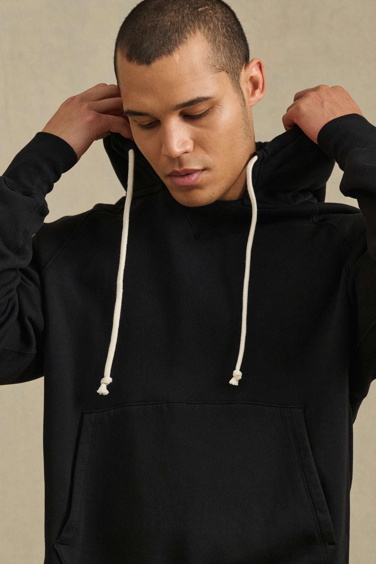 Wearing the BRODIE HOODIE by Velvet by Graham & Spencer, a person in a relaxed-fit black hoodie with white drawstrings adjusts the hood while looking down.